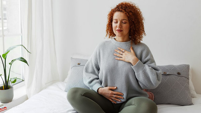 Portrait of plus-size curly redhead female in casual clothes doing pranayama technique sitting on bed next to window with closed eyes, putting hands on chest, breathing slowly and deeply Portrait of plus-size curly redhead female in casual clothes doing pranayama technique sitting on bed next to window with closed eyes, putting hands on chest, breathing slowly and deeply
