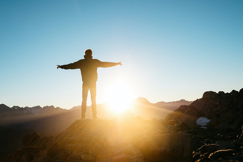 image-asset A Man standing with his arm open on the top of a mountain at the sunset
